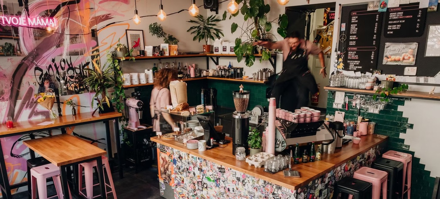 The colorful interior of Pinkiez Espresso Bar with a neon sign, graffiti mural, pink stools, and a barista in motion behind the sticker covered counter